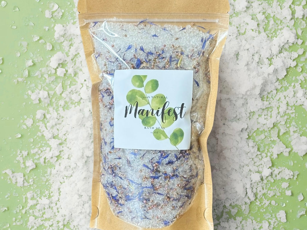 Manifest Bath Soak in a clear package with a botanical label, surrounded by bath salt on a green background.