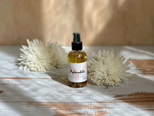 Aphrodite Goddess Ritual Oil in a spray bottle surrounded by soft white flowers on a rustic wooden surface.