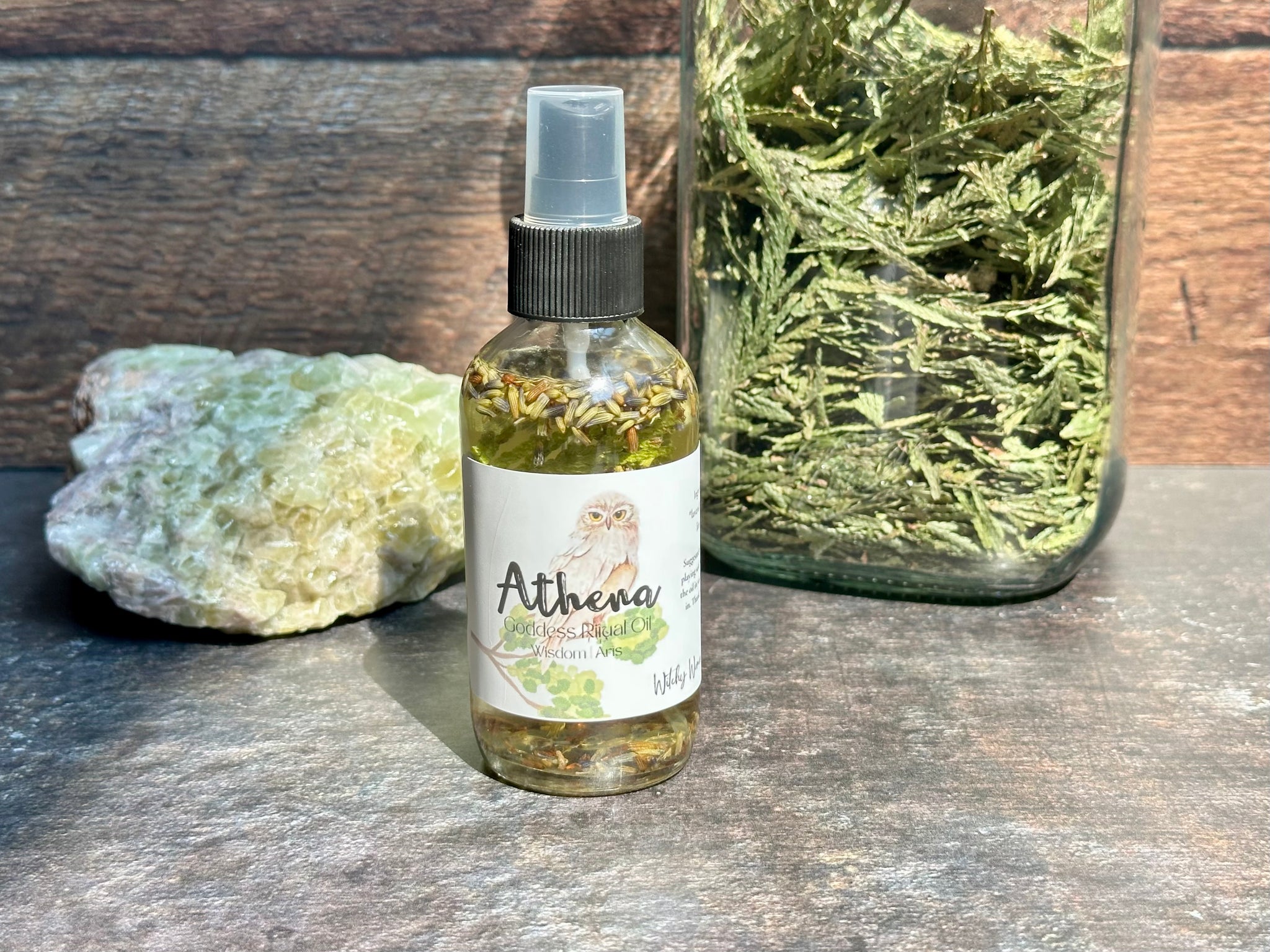 Bottle of Athena oil spray on a wooden surface with a jar of dried herbs and a rock.