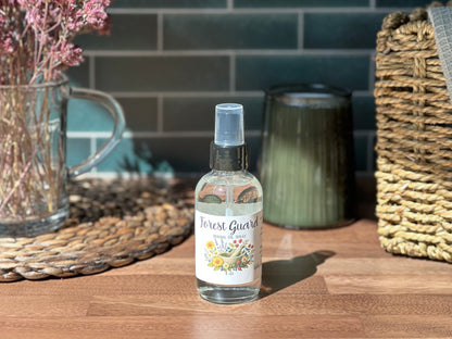 Forest Guard Herbal Oil Spray bottle on a wooden table with herbs and decorative items, natural bug repellant.
