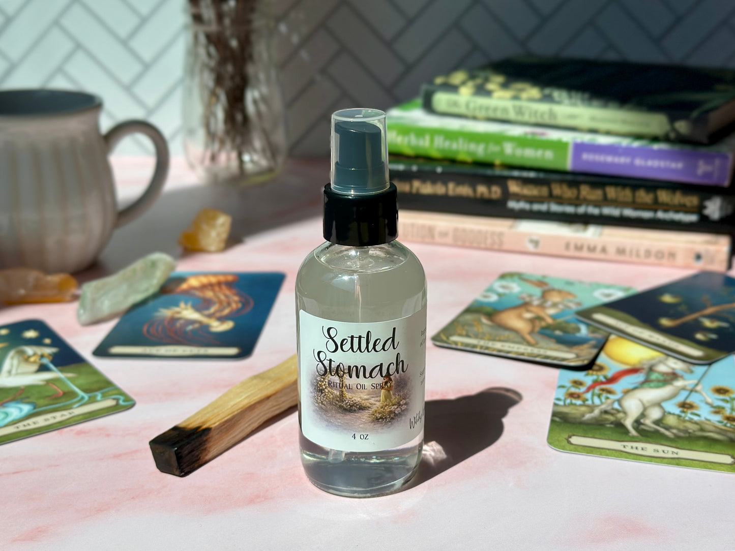 Settled Stomach Ritual Oil Spray bottle surrounded by books and tarot cards on a pink surface.