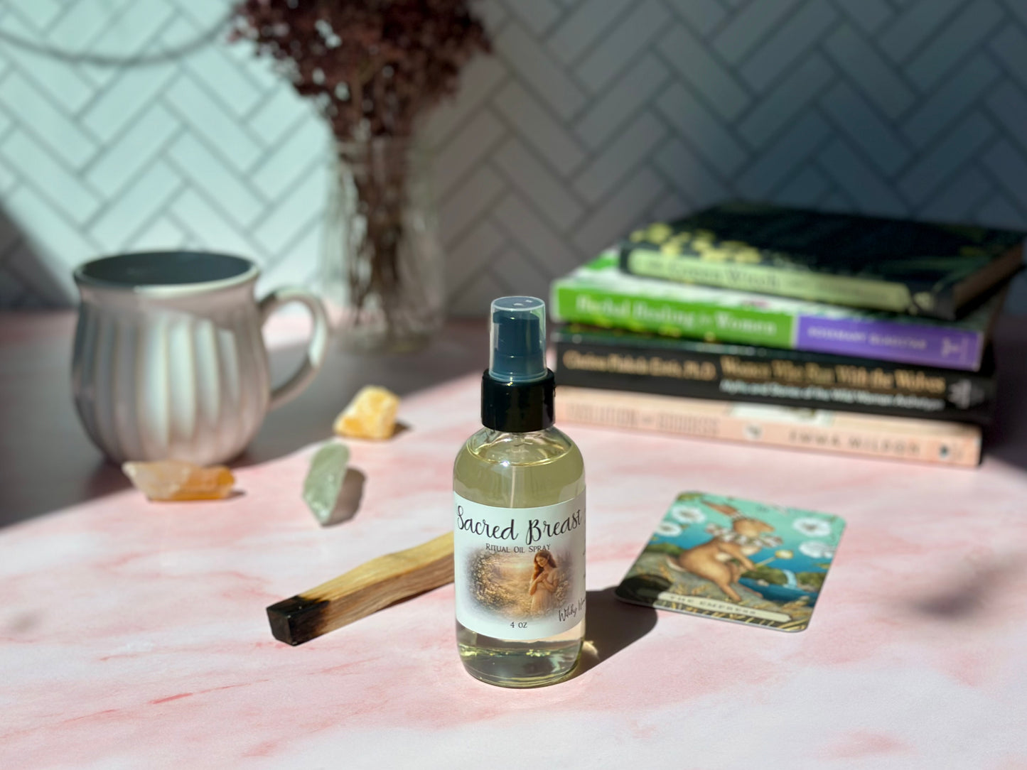 Sacred Breast Ritual Oil Spray bottle on a pink surface with books and crystals in a cozy setting.