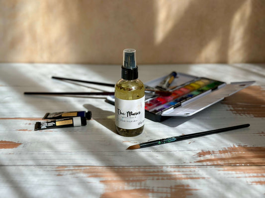 The Muses Goddess Ritual Oil bottle surrounded by paint tubes and brushes on a wooden surface.