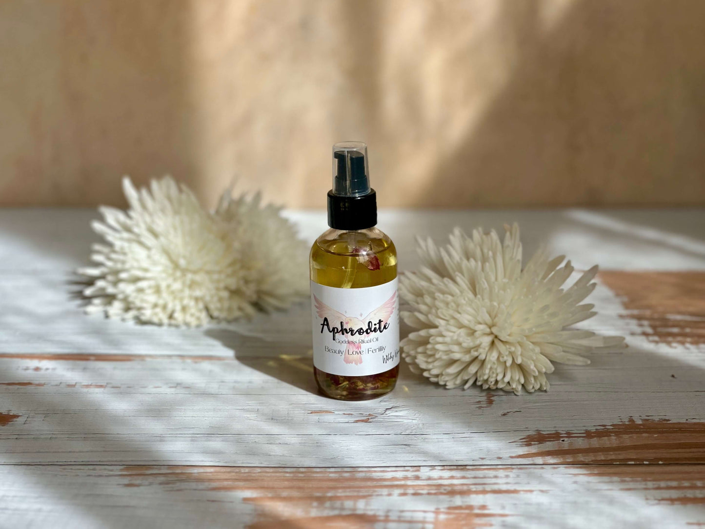 Aphrodite Goddess Ritual Oil in a spray bottle surrounded by soft white flowers on a rustic wooden surface.