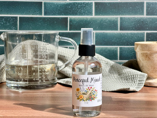 Bottle of 'Peaceful Mind' essential oil spray on a wooden surface with a measuring cup and towel in the background.