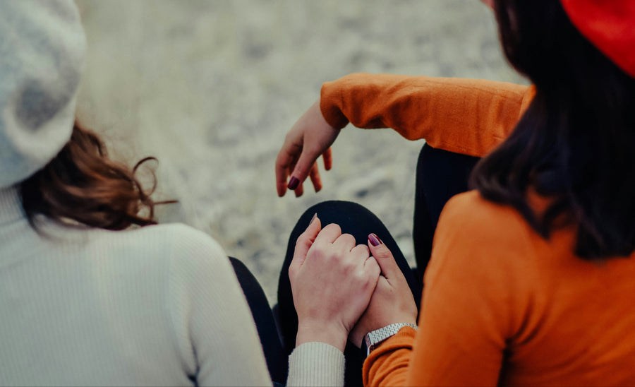 Two women sitting outdoors on a sandy surface, holding hands. One wears a white ribbed sweater and beanie, and the other wears a bright orange long-sleeve top and a red beret.
