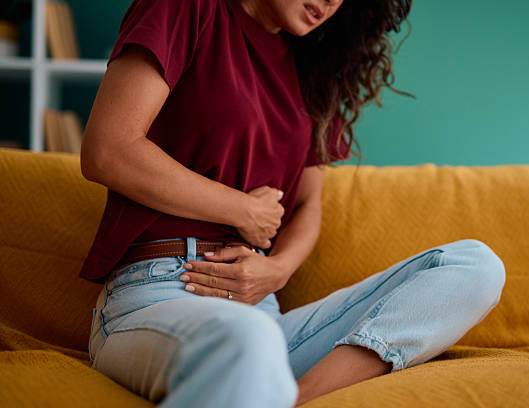 A woman sitting on a mustard yellow couch, clutching her stomach in discomfort. She is wearing a dark red t-shirt and light blue jeans, with a teal wall in the background.